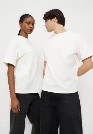 Două persoane îmbrăcate în tricouri albe simple, supradimensionate și pantaloni negri, stând în picioare pe un fundal alb.