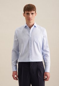 Camicia a maniche lunghe azzurro chiaro realizzata in tessuto morbido, con chiusura a bottoni nella parte anteriore, taschino sul petto, abbinata a pantaloni a pieghe scuri.
