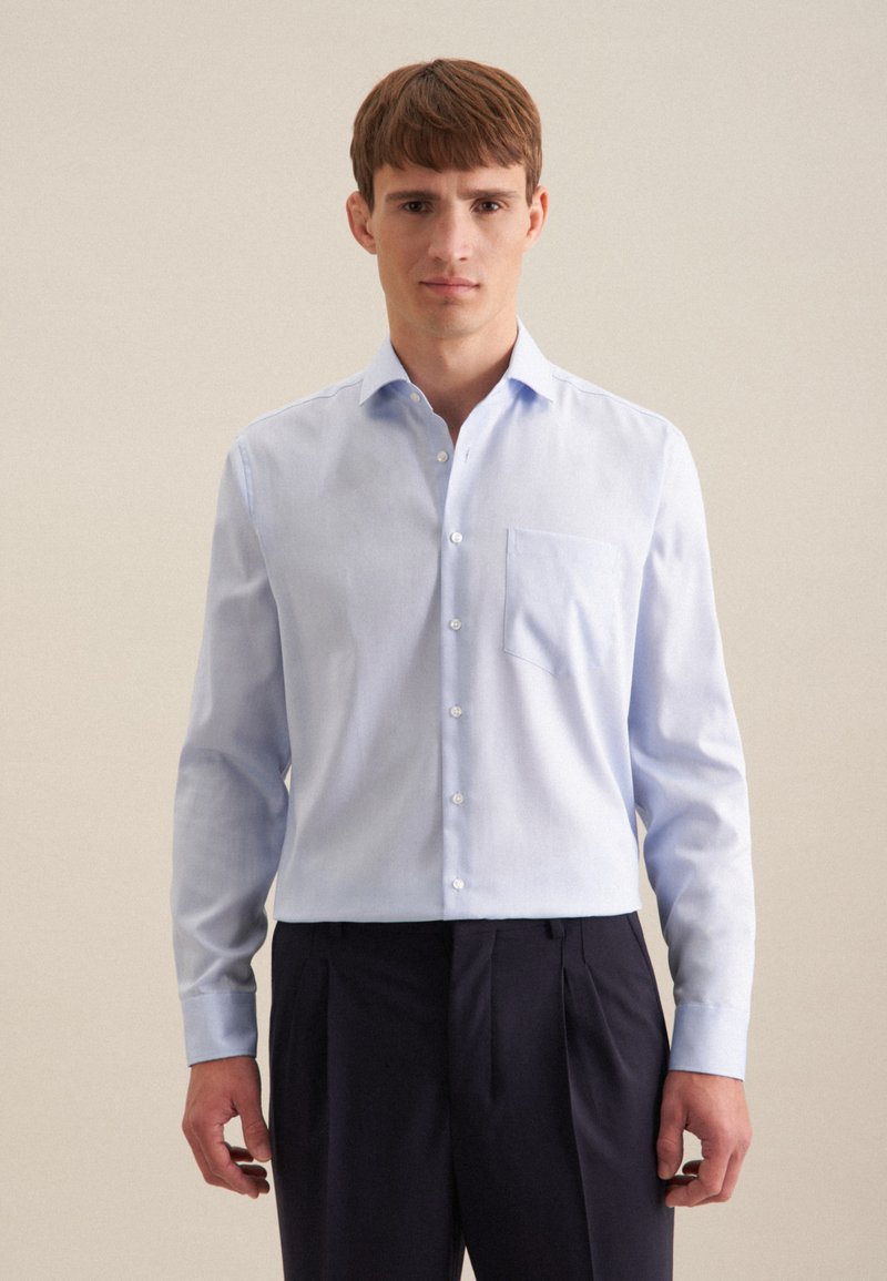 Camicia a maniche lunghe azzurro chiaro realizzata in tessuto morbido, con chiusura a bottoni nella parte anteriore, taschino sul petto, abbinata a pantaloni a pieghe scuri.