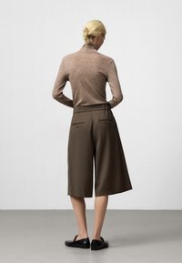 Brązowy długi sweter z wysokim kołnierzem, w parze z oliwkowymi szortami w szerokim kroju, mającymi dwie tylne kieszenie. Czarne buty wsuwane.