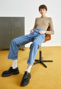 Beige gerippter Langarmshirt, hochtaillierte blaue Jeans, schwarze Plateauschuhe, sitzend auf einem braunen Stuhl auf einem gelben Boden.