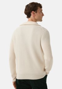 Mann trägt einen cremefarbenen, gerippten Strickpullover mit Schalkragen, von hinten gezeigt und leicht nach rechts gewandt.