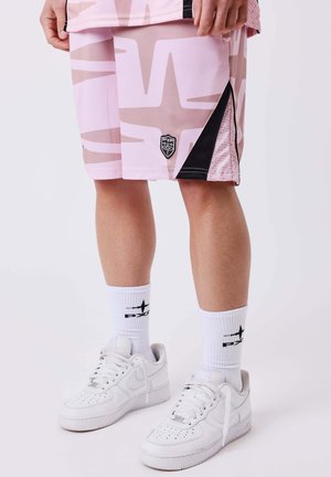 Persoon die roze en beige korte broek met patroon draagt met zwarte details, witte sokken met logo en witte sneakers met veters, staand op een witte achtergrond.