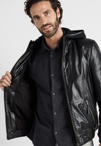 Schwarze Lederjacke mit Reißverschluss und Kapuze, offen präsentiert. Zu den Merkmalen gehören seitliche Taschen und eine glatte Oberfläche, getragen über einem dunklen Hemd.