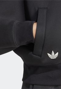 Mano nella tasca di una felpa nera con polsini a coste e logo Adidas Trefoil bianco vicino all'orlo.