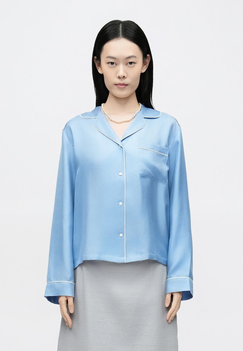 Világoskék szatén pizsamaing fehér paszpól díszítéssel, hosszú ujjú, mellzsebbel és gombos elülső résszel. Puha tapintású és kényelmes szabású.