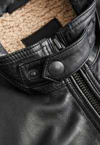 Cuello de chaqueta de cuero negro con un forro suave y texturizado en color beige, un botón a presión con logo y un cierre de cremallera de metal.