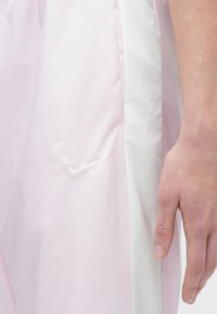 Χέρι να ακουμπά δίπλα σε ανοιχτό ροζ και λευκά κοντά παντελόνια με μικρό λευκό λογότυπο Nike κοντά στην περιοχή της τσέπης.