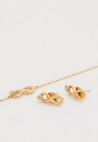 Σετ χρυσών κοσμημάτων με σκουλαρίκια σε σχήμα άπειρου και αντίστοιχο βραχιόλι. Διακοσμημένο με μικρές διάφανες πολύτιμες πέτρες, παρουσιάζοντας λεία υφή.