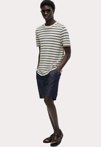 Camiseta de manga corta a rayas en crema y azul marino, combinada con pantalones cortos de mezclilla oscura. Usada con sandalias marrones, con un ajuste relajado.