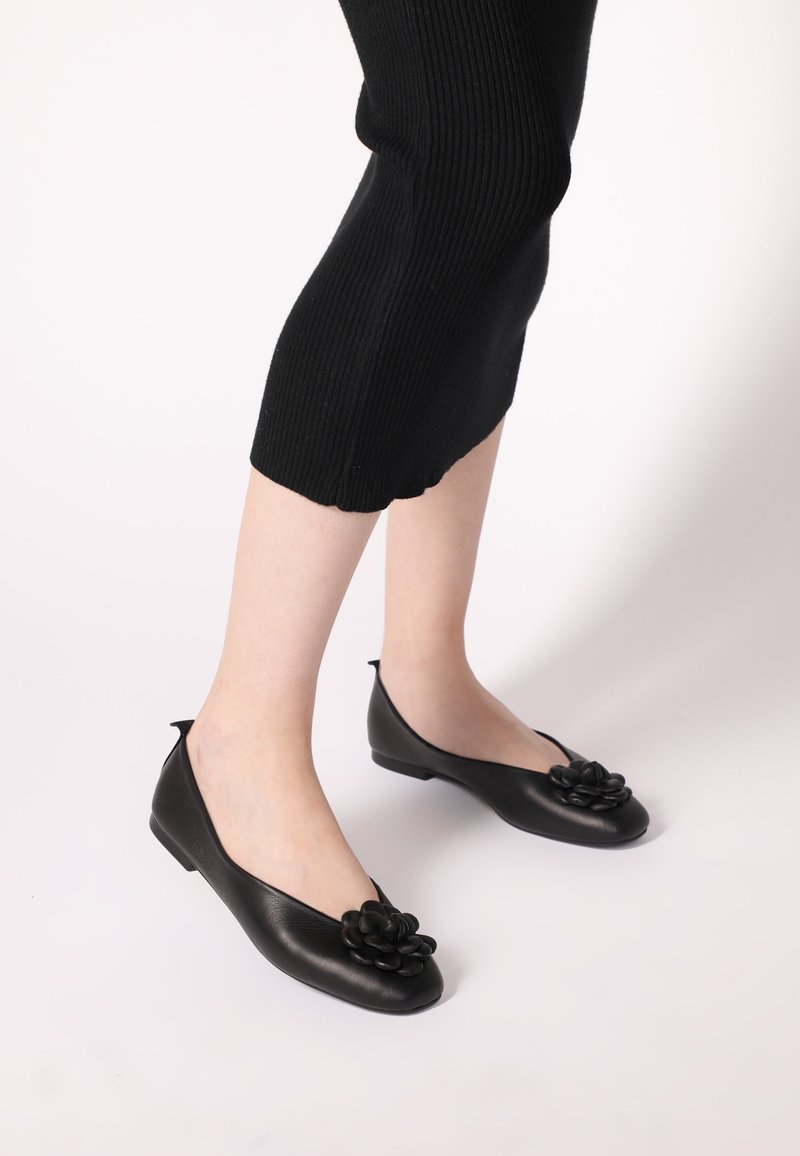 Schwarze Leder-Ballettschuhe mit einer Blumenapplikation an der Zehe, runder Form und glatter Textur. Kombiniert mit schwarzen, gerippten, knöchellangen Hosen.