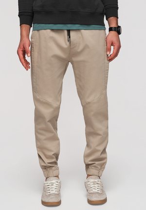 Man draagt beige joggerbroek, grijze sneakers, een zwarte horloge, een groen hemd en een zwart sweatshirt, staand tegen een effen achtergrond.