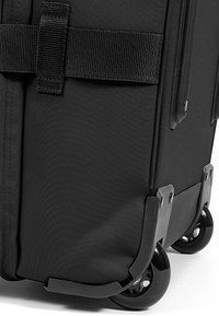 Valigia nera a rotelle con un esterno in tessuto resistente, angoli rinforzati e ruote nere scorrevoli. Include una robusta cinghia in nylon.