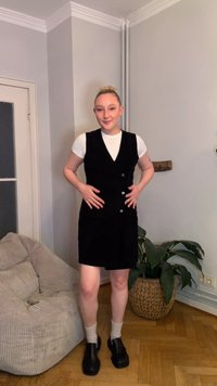 Crna kordirana haljina s V-izrezom i gumbima, nošena preko bijele majice, uparena s crnim cipelama i bež čarapama; unutarnji prostor s biljkama.