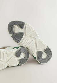 Podplati belih in sivih superg z teksturiranim vzorcem teksta v geometrijskih oblikah na svetlem ozadju.