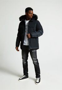 Čierna prešívaná bunda s kapucňou podšitou kožušinou, so zipsom a viacerými prednými vreckami. Nosená s poškodenými tmavými džínsami a čiernymi teniskami.