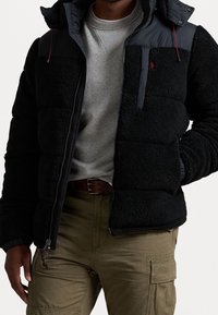 Man med svart puffjacka över grå tröja och olivgröna cargobyxor med brunt bälte, händerna vilande vid höfterna.