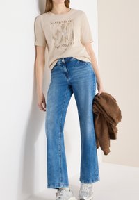 Femme portant un T-shirt graphique beige et un jean bleu, tenant une veste en polaire marron, debout devant un mur bicolore beige et blanc.