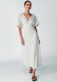 White maxi dress with black polka dots, V-neckline, short voluminous sleeves, and a flowing texture. Paired with black strappy sandals.