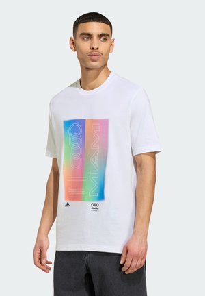 Mann trägt weißes T-Shirt mit buntem vertikalem Schriftzug "1000 MIAMI" und Logos, kombiniert mit einer dunklen Hose, steht vor hellem Hintergrund.