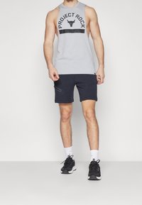 Camiseta sin mangas de color gris claro con gráfico "PROJECT ROCK" y acentos negros, combinada con pantalones cortos azul marino con bolsillos laterales y una textura suave.