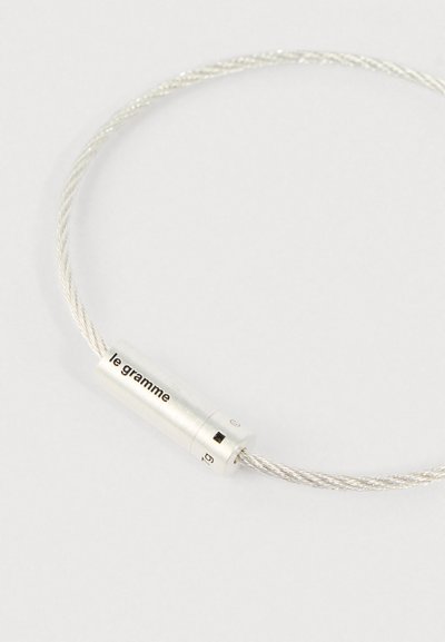 Srebrna uvrta žica narukvica s cilindričnom kopčom na kojoj je ugravirano "le gramme" i "1g" na svijetloj pozadini.