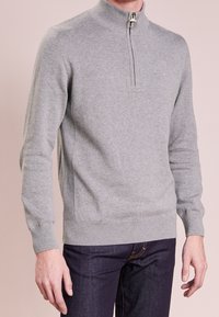 Mann trägt einen hellgrauen Langarmpullover mit einem Viertel-Reißverschluss sowie gerippten Bündchen und Saum, kombiniert mit dunkelblauen Jeans.