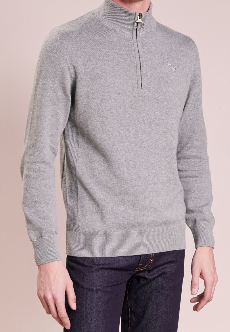 Mann trägt einen hellgrauen Langarmpullover mit einem Viertel-Reißverschluss sowie gerippten Bündchen und Saum, kombiniert mit dunkelblauen Jeans.