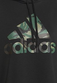 Hoodie noir avec un grand logo Adidas en camouflage vert. Le design comprend une finition texturée avec des cordons de serrage et des embouts en métal.