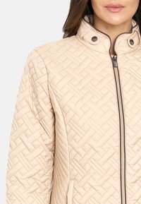 Chaqueta acolchada beige claro con un alto cuello, cierre frontal y cierres a presión. Presenta patrones diagonales texturizados.