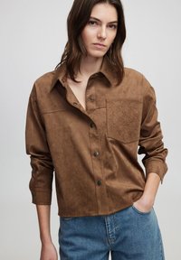 Bruine faux-suède shirt met een knoopsluiting aan de voorkant, een borstzak en een tonale textuurpatroon op de zak. Ronde hals en relaxte pasvorm.