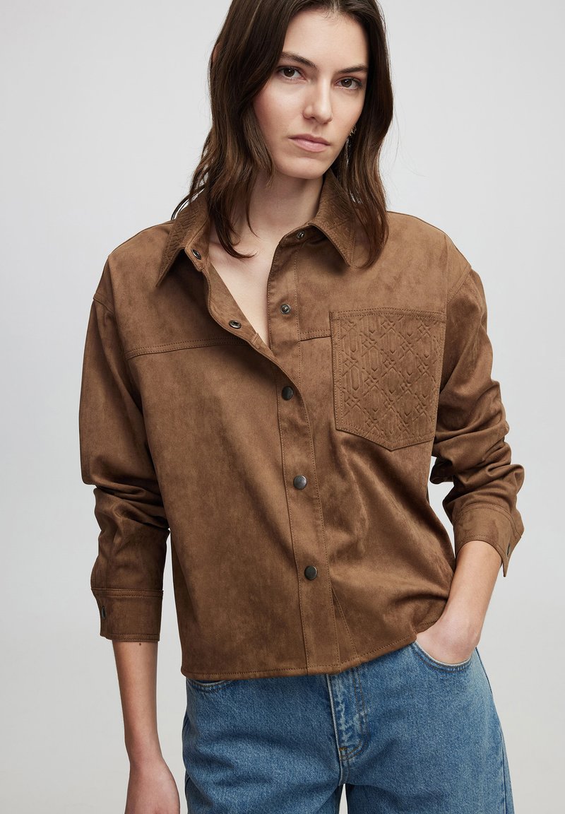 Bruine faux-suède shirt met een knoopsluiting aan de voorkant, een borstzak en een tonale textuurpatroon op de zak. Ronde hals en relaxte pasvorm.