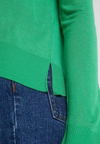 Maglione verde in maglia con polsini e orlo a coste, con fessure laterali e una texture liscia, abbinato a jeans blu scuro.