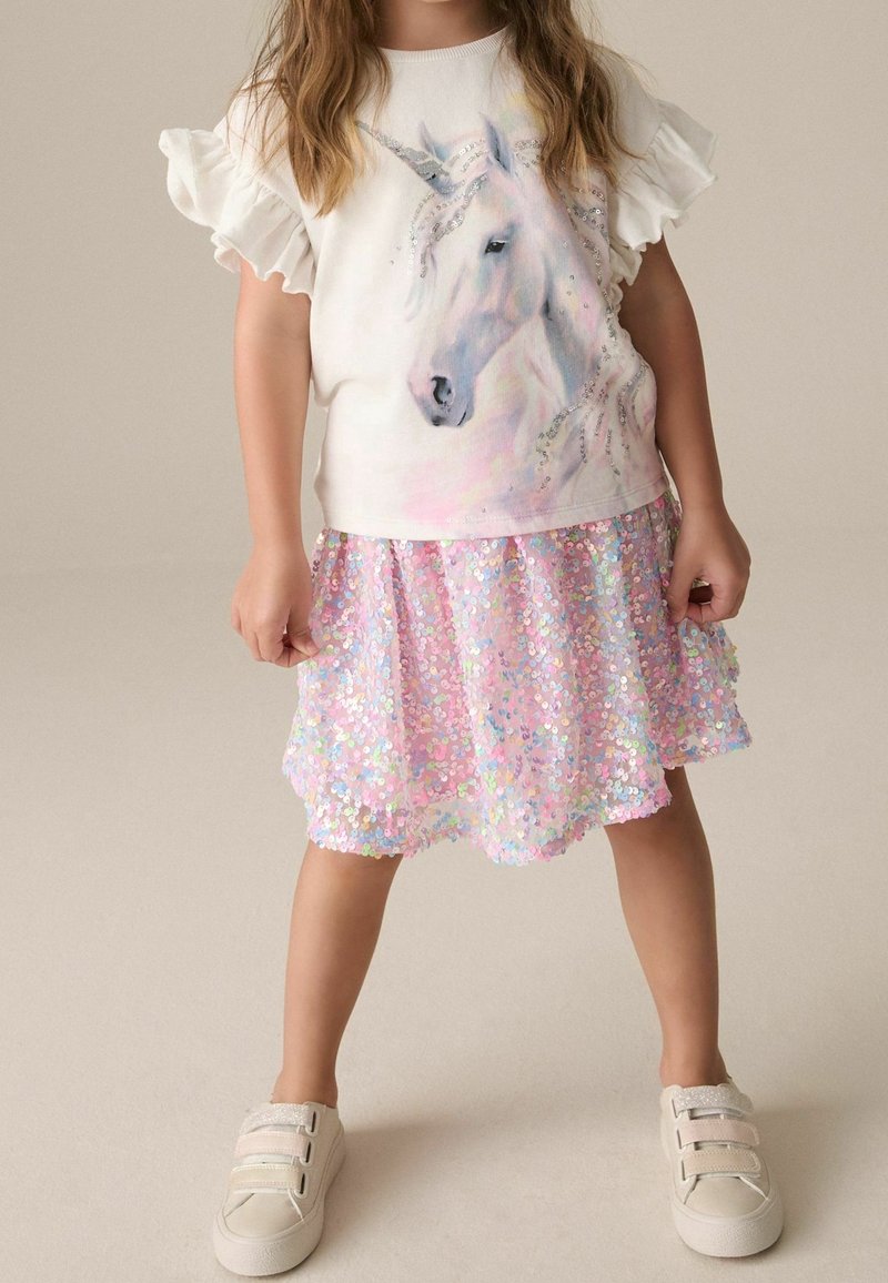 Enfant portant une chemise blanche avec un motif de licorne, une jupe rose à paillettes, et des baskets blanches à scratch.