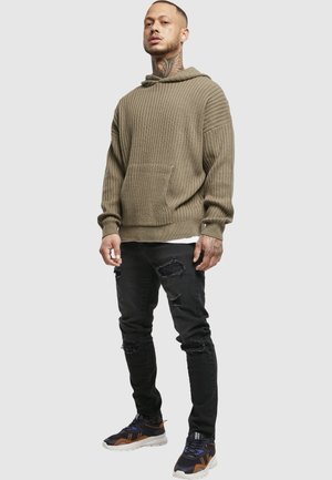 Homme avec des tatouages au cou et aux mains, portant un pull côtelé marron, un jean noir déchiré et des baskets noires sur un fond gris clair.