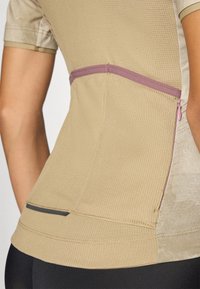 Beige treningsskjorte med teksturert stoff, kontrasterende rosa glidelåslomme og svarte detaljer i kanten. Korte ermer, tettsittende design.