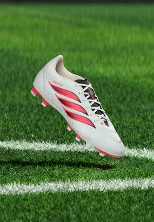 Chaussure de football Adidas Predator blanche et rose avec des détails noirs flottant au-dessus de l'herbe verte près d'une ligne blanche du terrain.