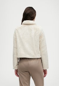 Kratka bež jakna od umjetnog krzna s mekanom teksturom, skraćenim dizajnom i preklopnim ovratnikom. Ima duge rukave i ležeran kroj.