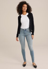 Svart stickad cardigan med knappar, vit t-shirt med rund hals, ljusblå skinny jeans och svarta spetsiga ballerinaskor. Modellen står mot en neutral bakgrund.