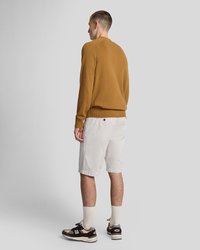 Mann, der mit dem Rücken zur Kamera steht, trägt einen braunen Pullover, hellgraue Shorts, cremefarbene Socken und braune sowie beige Turnschuhe vor einem schlichten grauen Hintergrund.