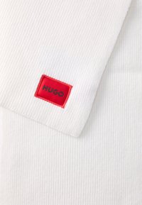 Witte geribbelde stoffen sjaal met een rood rechthoekig label waarop de tekst "HUGO" in zwart staat. De textuur lijkt zacht en glad te zijn.