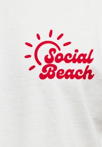 Weißes Baumwoll-T-Shirt mit rotem Text, der "Social Beach" liest, umgeben von Sonnenstrahlen-Grafiken.