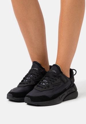 Jambes nues portant des baskets Diesel noires avec semelles épaisses et lacets noirs sur fond blanc.