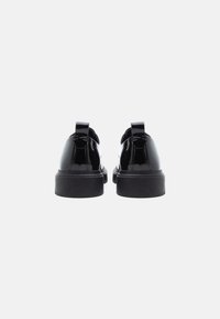 Um par de sapatos plataforma de couro preto brilhante, vistos de trás, sobre um fundo branco liso.