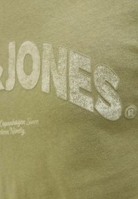 Olijfgroene t-shirt met een getextureerde witte print van "JONES" en kleinere tekst daaronder. Zachte stof met een subtiel textuurpatroon.