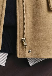 Tan wool coat with a visible gold metal zipper, featuring a smooth texture and a side snap detail; layered over a dark knit and striped shirt.