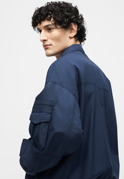 Νεαρός άνδρας με σγουρά σκούρα μαλλιά, φορώντας σκούρο μπλε μπουφάν με μεγάλη τσέπη στο μανίκι, κοιτάζει πάνω από τον ώμο του.