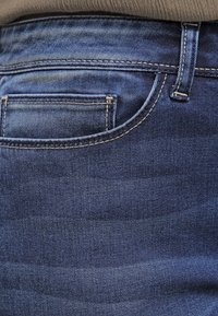 Tummansininen farkkukangas sisältää sileän pinnan, jossa on hienovaraisia kulutuksen jälkiä, viiden taskun muotoilu ja kontrastiväriset keltaiset ompeleet saumoissa.