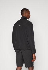 Svart vindjakke med høy krage, elastiske mansjetter og en diskret logo på baksiden. Kombinert med matchende svarte shorts.