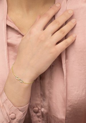Pulsera de cadena de oro con un acento de símbolo de infinito. La pulsera presenta un diseño delicado, complementado por una blusa de seda de color rosa claro.